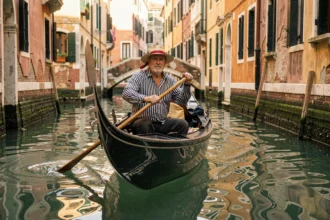 Gondolier en tenue traditionnelle pilotant une gondole dans un canal étroit de Venise avec des façades de palais colorées en arrière-plan